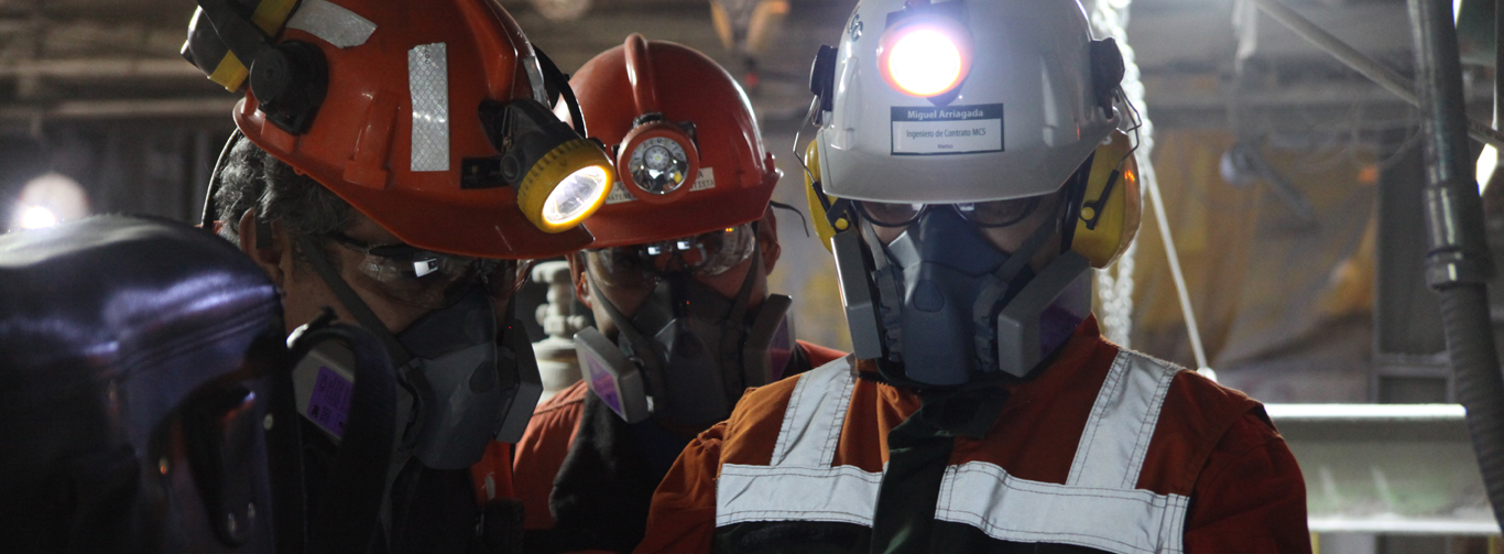 Three mining engineers with safety gear on looking down at one person's hands.