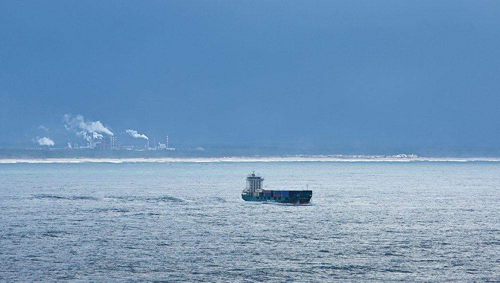 A ship with cargo pictured in the middle of the sea with coast in the far horizon.